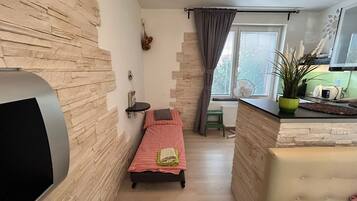 Una habitación pequeña con una cama, una mesita, una silla, un ventanal con cortinas y una planta en maceta.