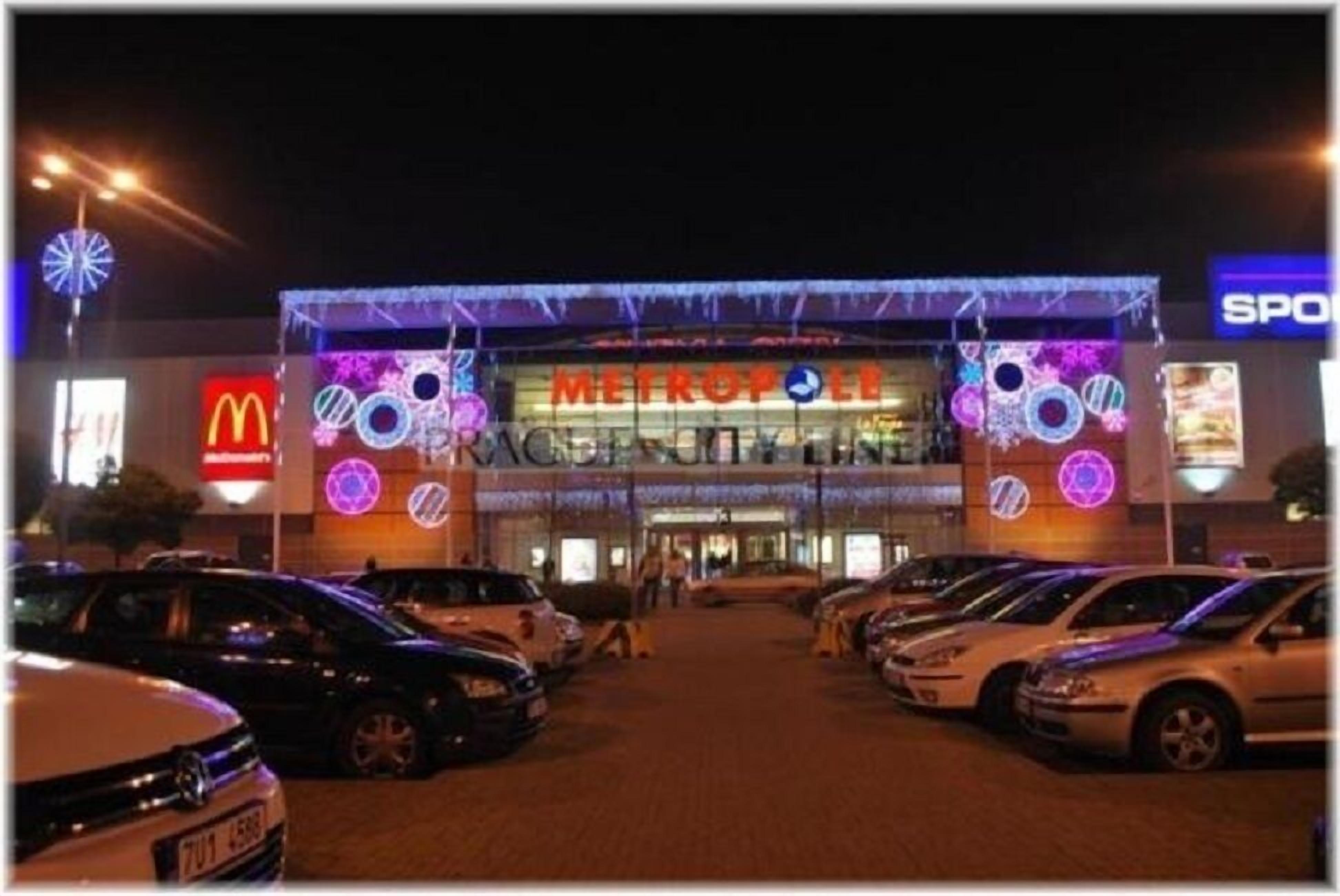 Un estacionamiento con autos y un edificio con letreros iluminados, incluyendo McDonald's y una tienda llamada METROPOLIS.