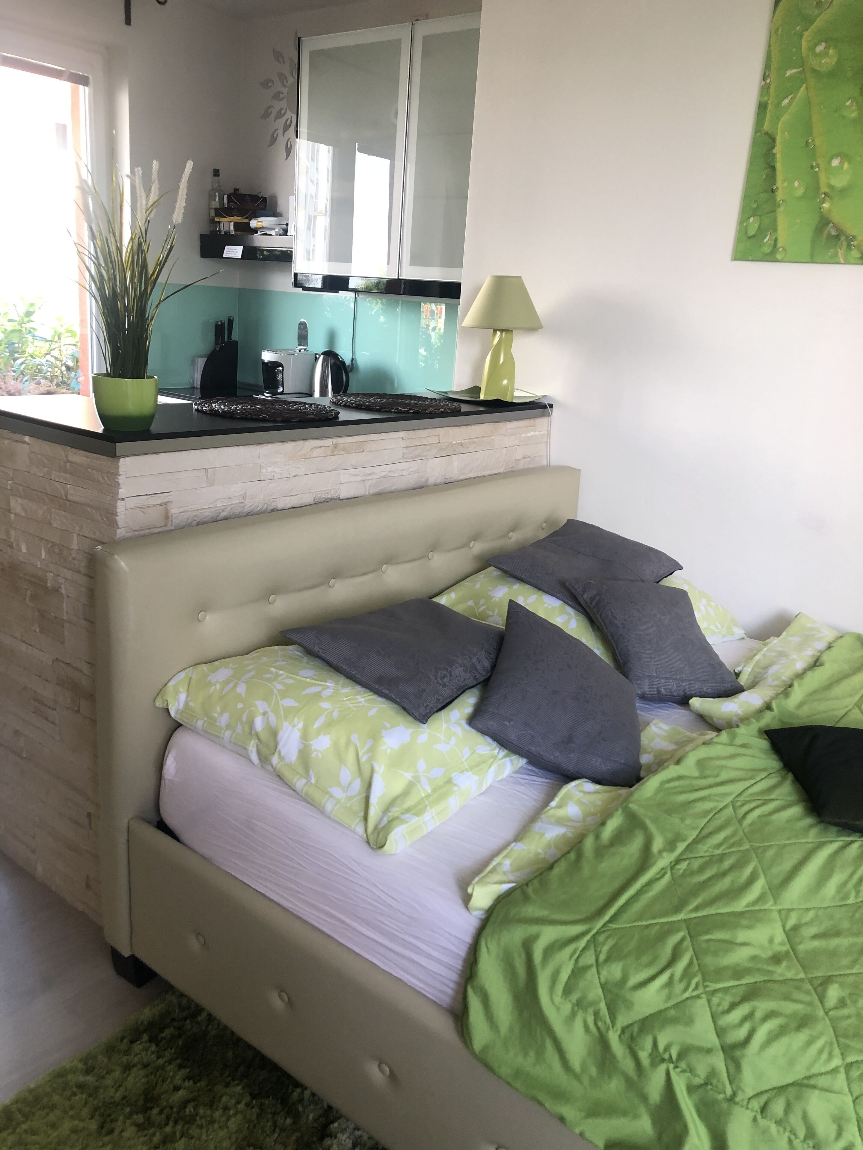 A bedroom with a bed, green and white bedding, a green rug, a small kitchen area with a stove, and a window.
