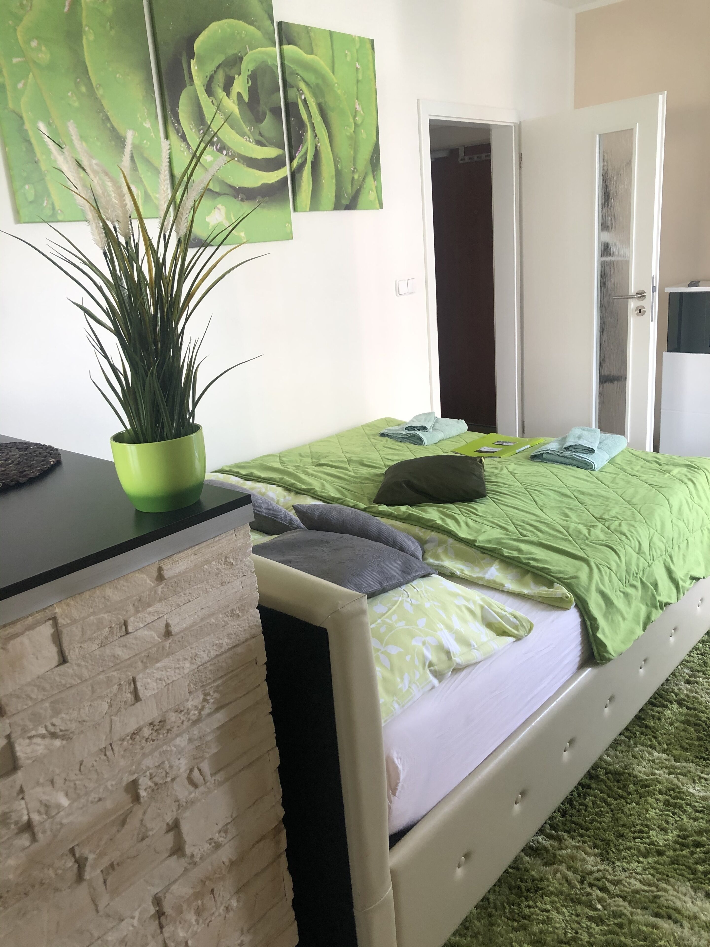 Ein Schlafzimmer mit grüner Bettdecke, einem Blumentopf, einem Schreibtisch und einem Kleiderschrank.