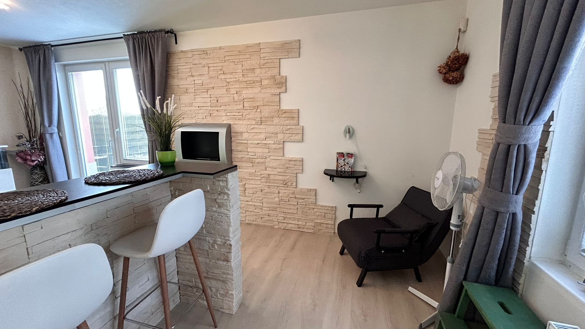 A compact living space with a kitchen area, a seating area with a chair and a small table, a television, and a fan.