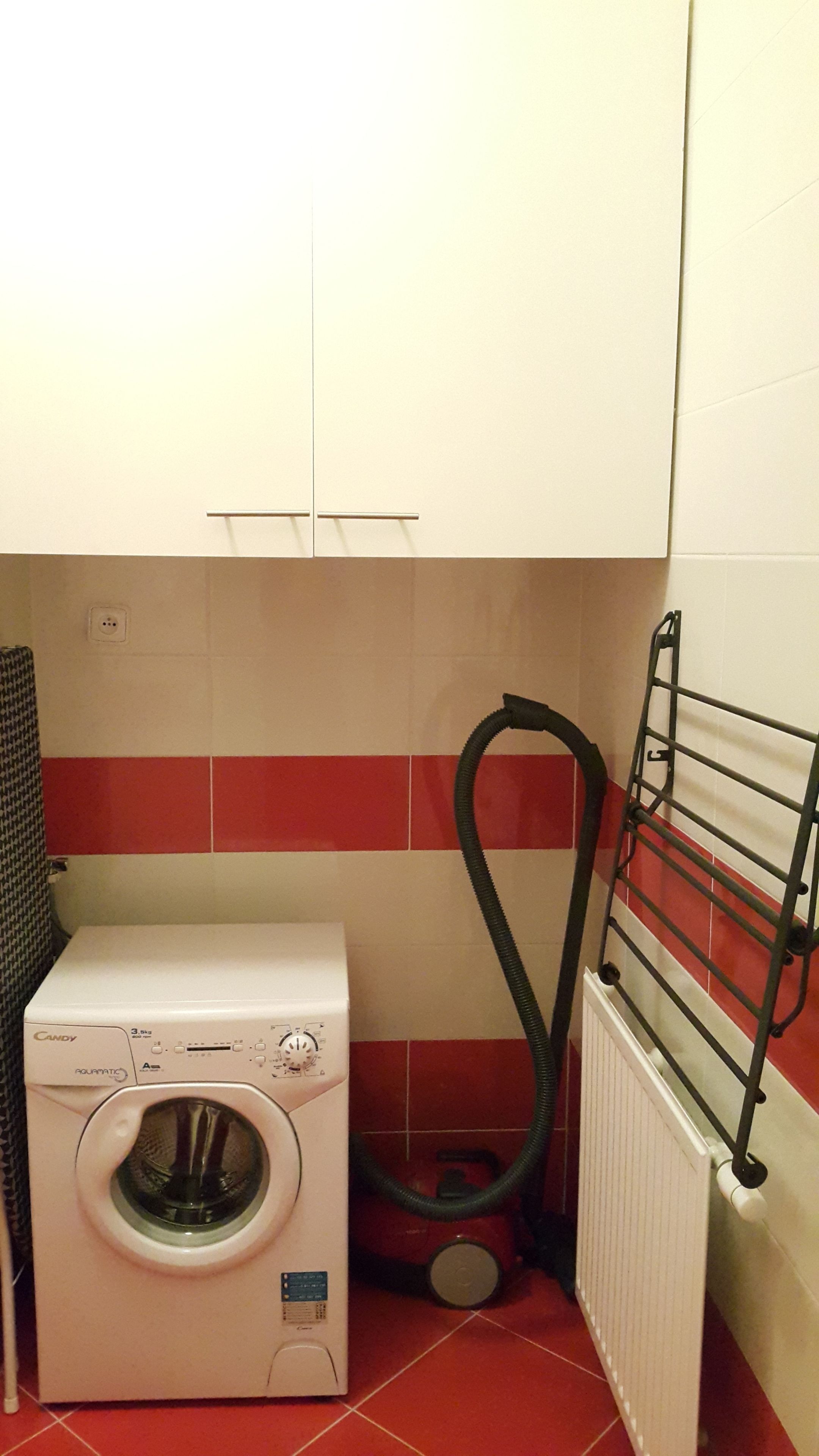 A compact laundry room with a front-loading washing machine, a hose, and a radiator.