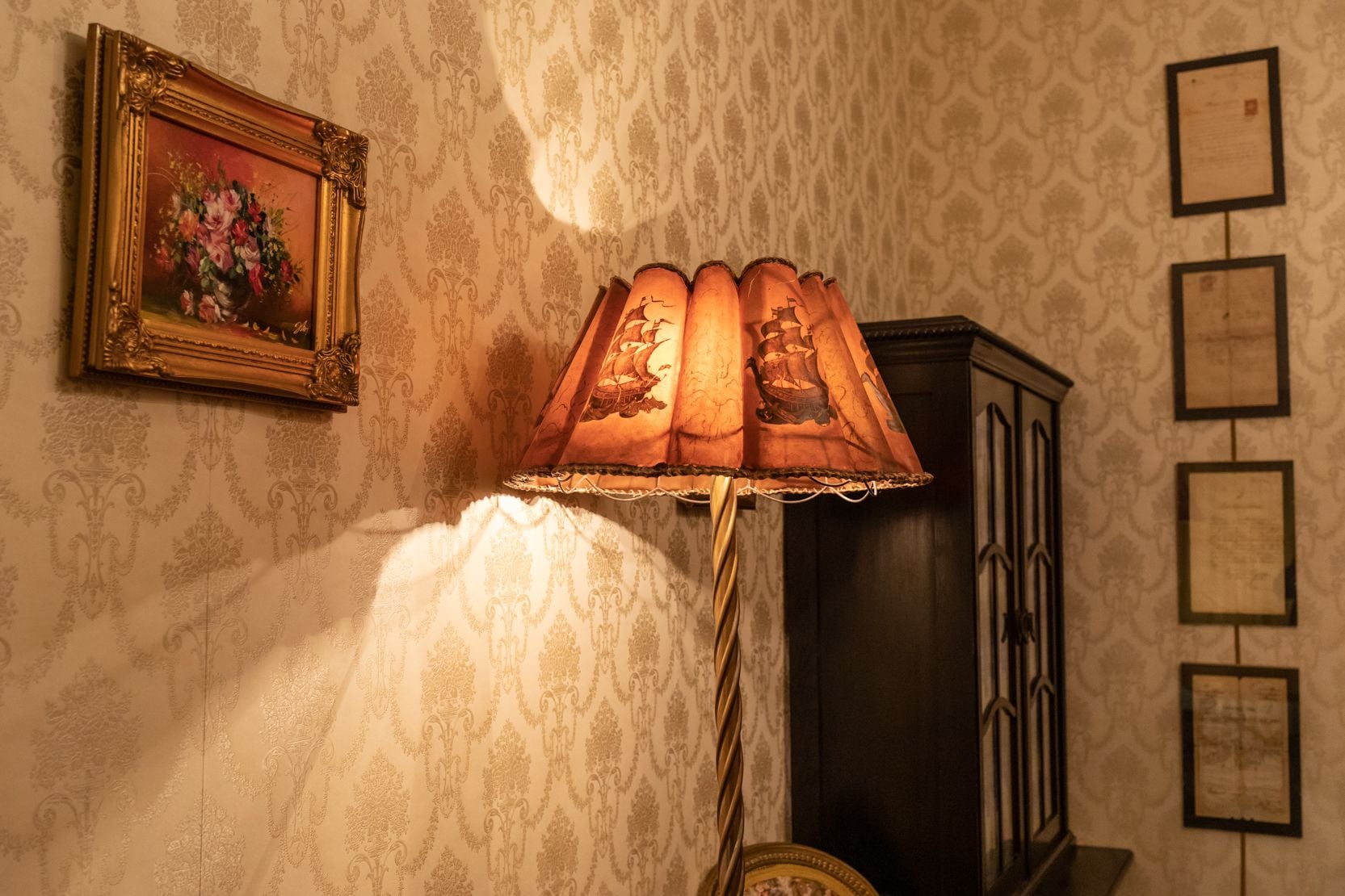 Een kamer met behang met patroon, een lijst met schilderij, een lamp met decoratieve kap en een kast.