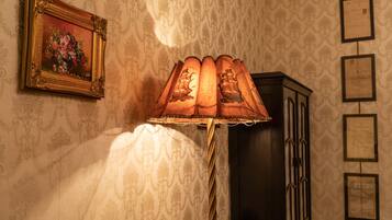 Een kamer met behang met patroon, een lijst met schilderij, een lamp met decoratieve kap en een kast.