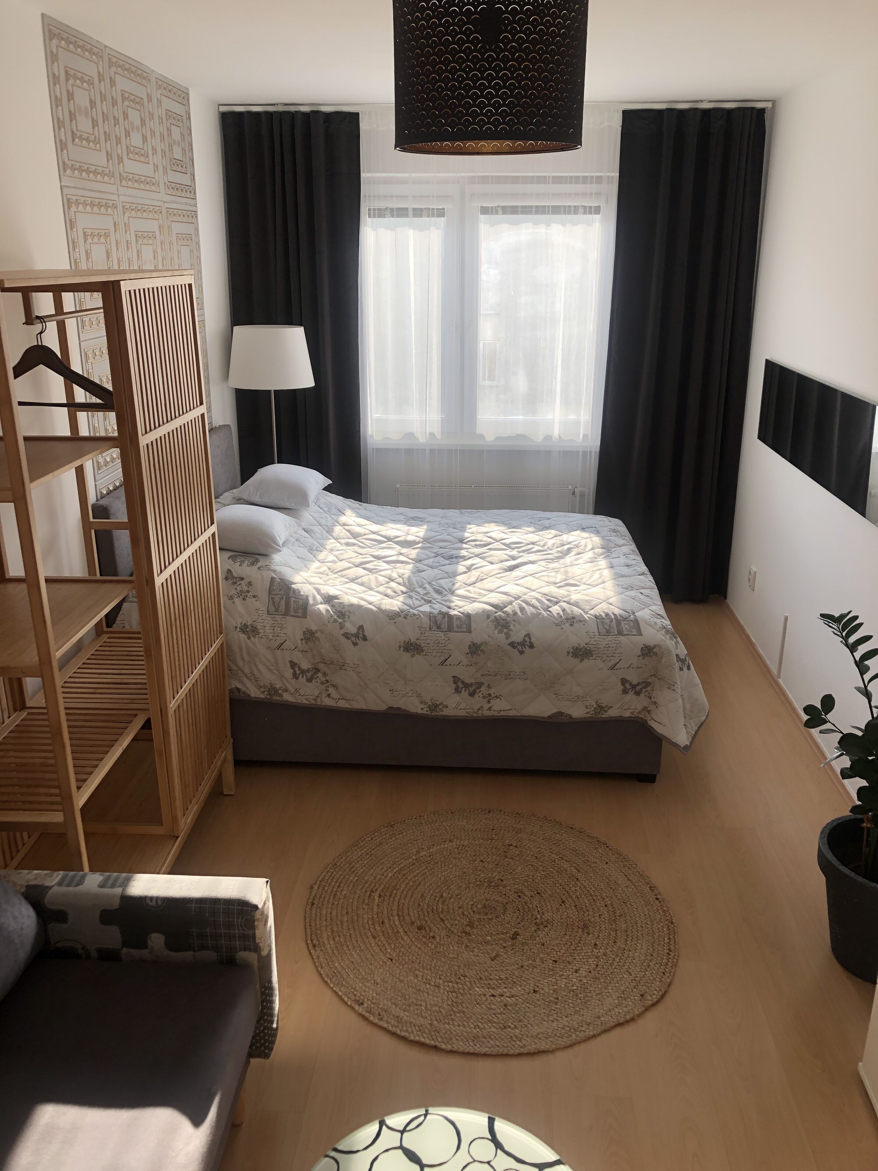 Ein Schlafzimmer mit einem Bett, einem Holzschrank, einem Nachttisch, einer Lampe, einem Fenster mit Vorhängen und einer Zimmerpflanze.