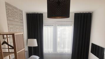 Ein Schlafzimmer mit einem Bett, einem Holzschrank, einem Nachttisch, einer Lampe, einem Fenster mit Vorhängen und einer Zimmerpflanze.