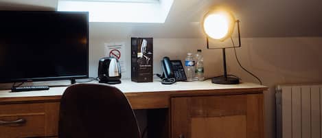 A bedroom with a skylight, a desk with a computer, a chair, a bed, and a bedside table with a lamp.