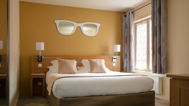 Una habitación de hotel con una cama grande, dos mesitas de noche, una lámpara de pared y un ventanal con cortinas.