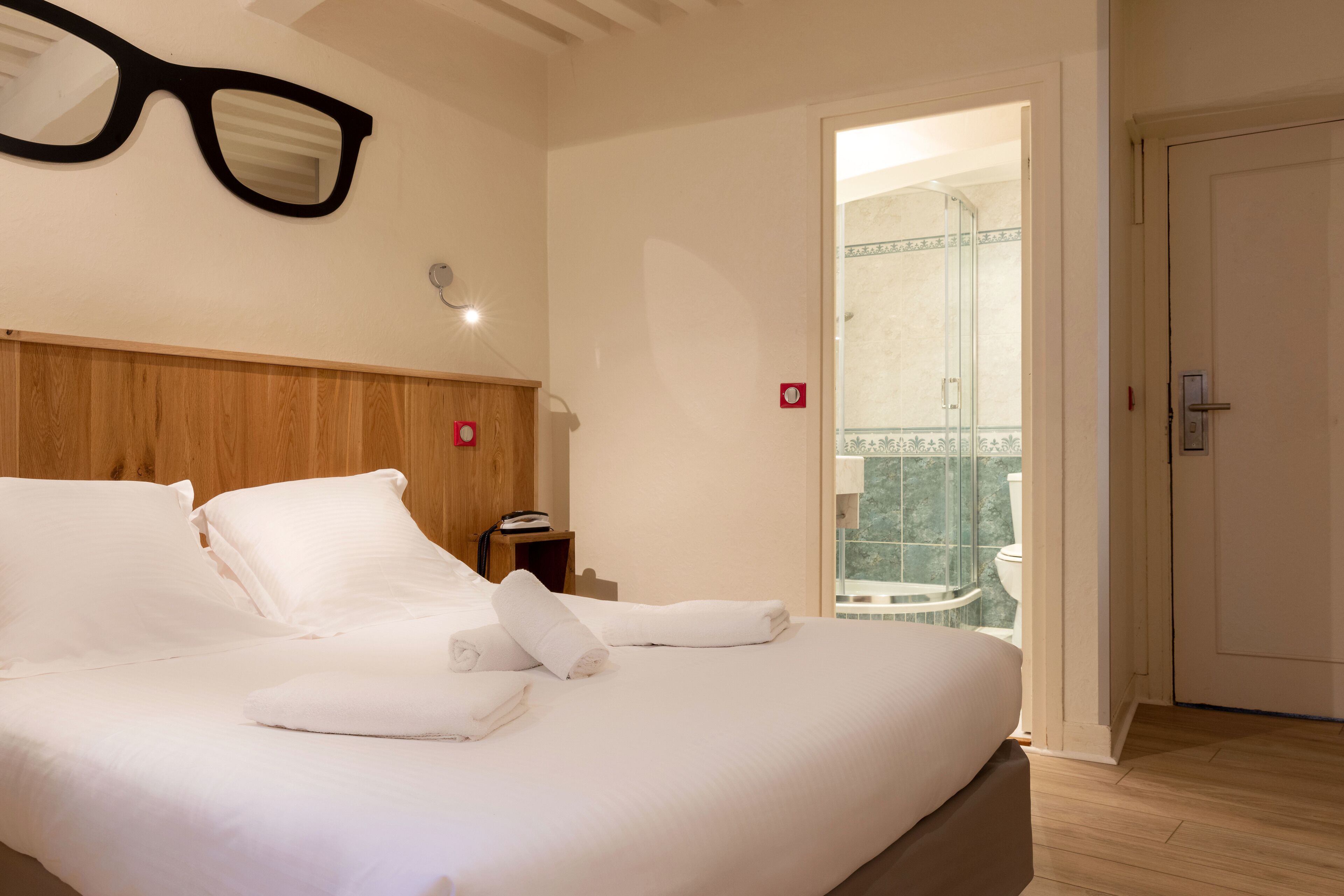 Habitación de hotel con cabecera de madera, cama con ropa blanca y baño a la vista a través de una puerta abierta.