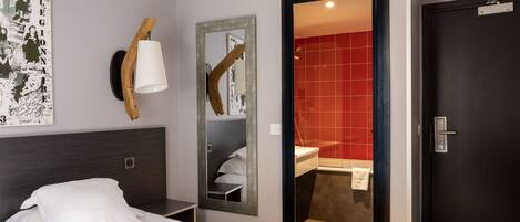 A hotel room with a bed, a mirror, a wall-mounted lamp, and a red-tiled bathroom visible through an open door.