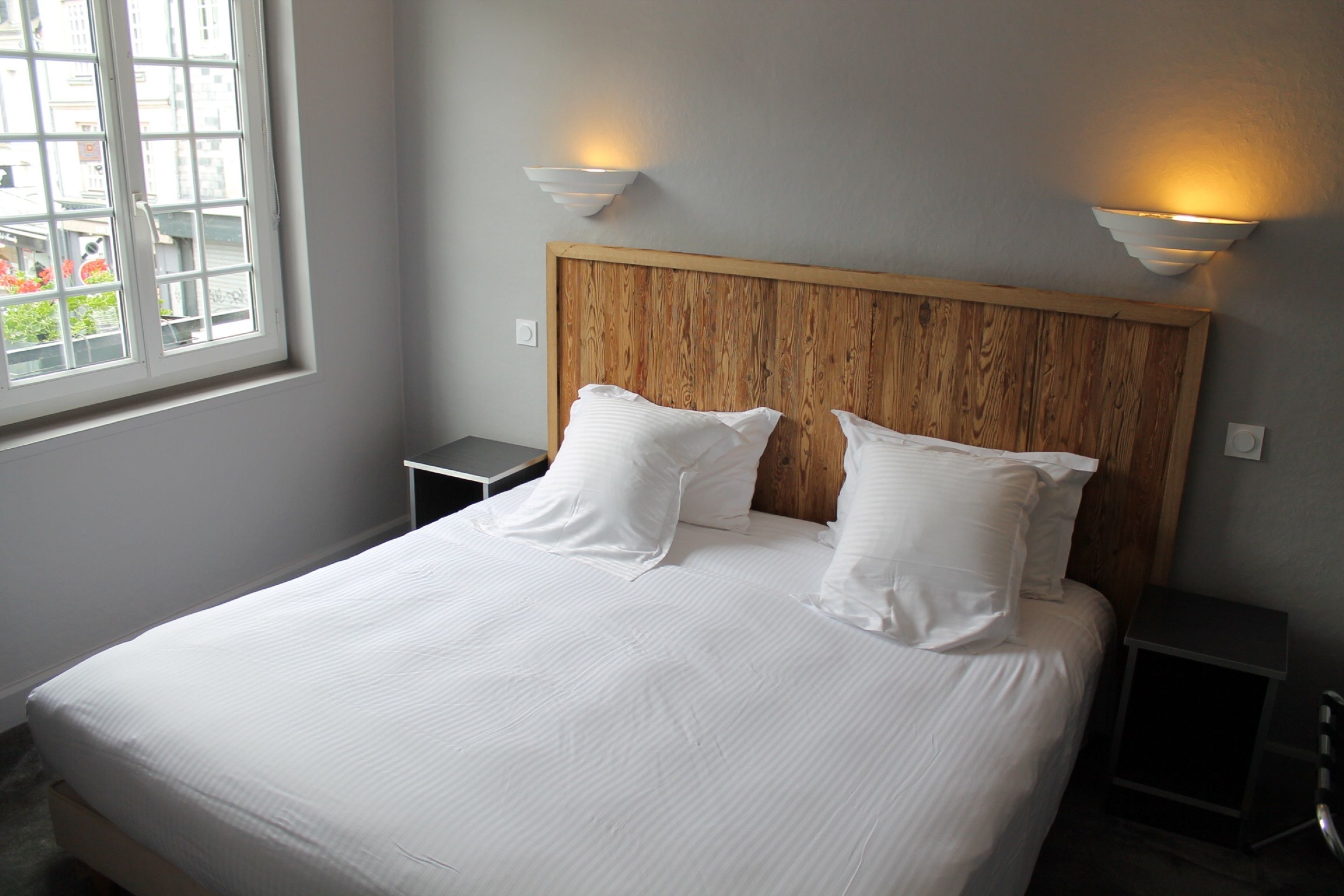 Una cama bien hecha con sábanas blancas y dos almohadas, un cabecero de madera y dos lámparas fijadas a la pared.
