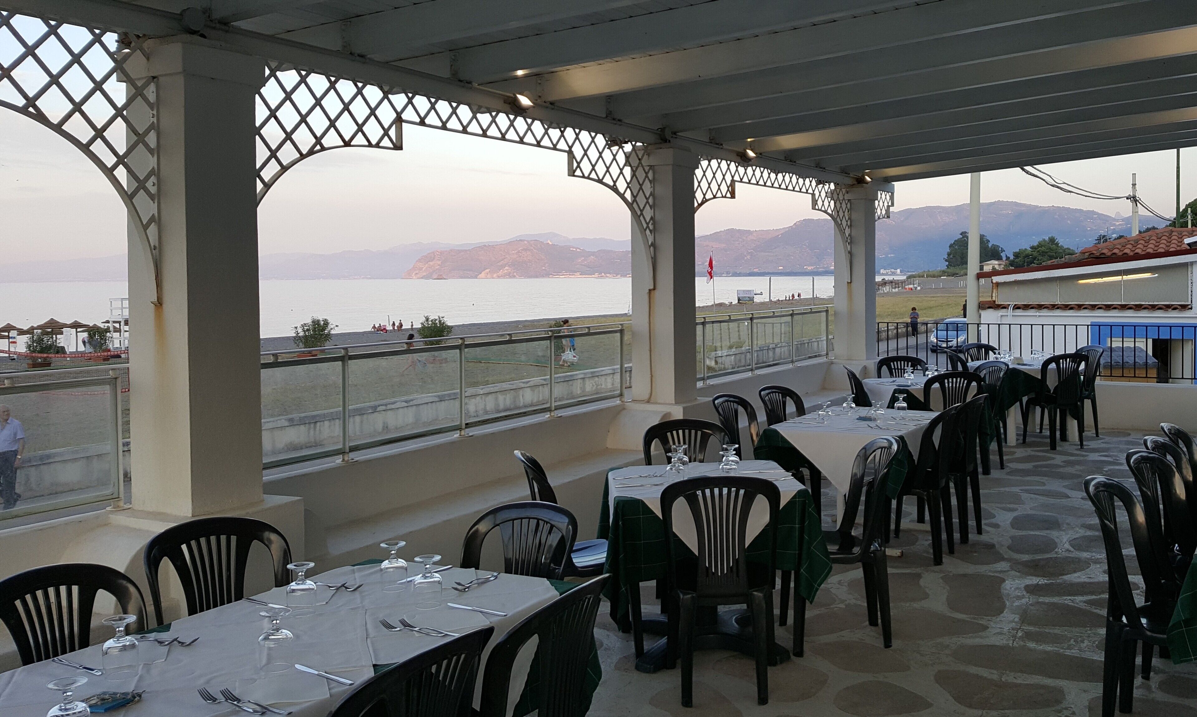Un área de comedor al aire libre con mesas listas para recibir clientes, con vistas a un cuerpo de agua y montañas distantes.