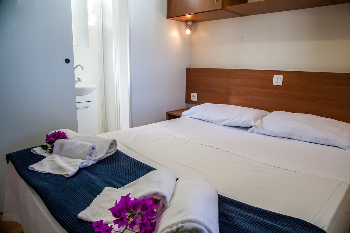 Een netjes opgemaakt bed met witte lakens en kussens, een houten hoofdbord en een klein nachtkastje met een lamp.