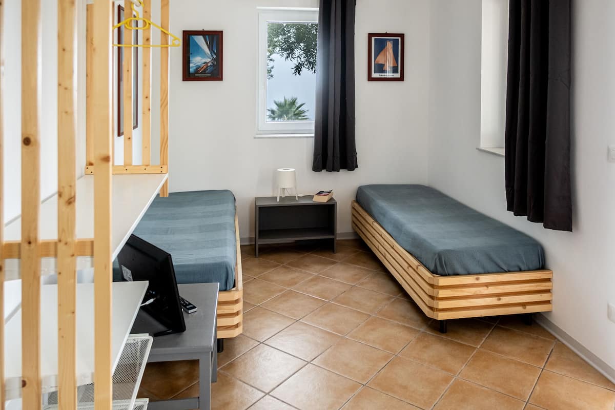 Een kleine, compacte kamer met twee houten stapelbedden, een bureau met een laptop, een klein nachtkastje en een raam met gordijnen.
