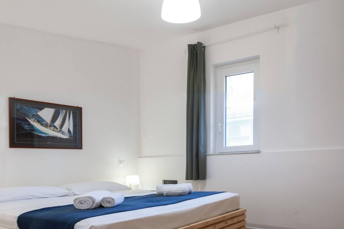 Een hotelkamer met een bed, een raam met gordijnen, een schilderij aan de muur en een lamp.