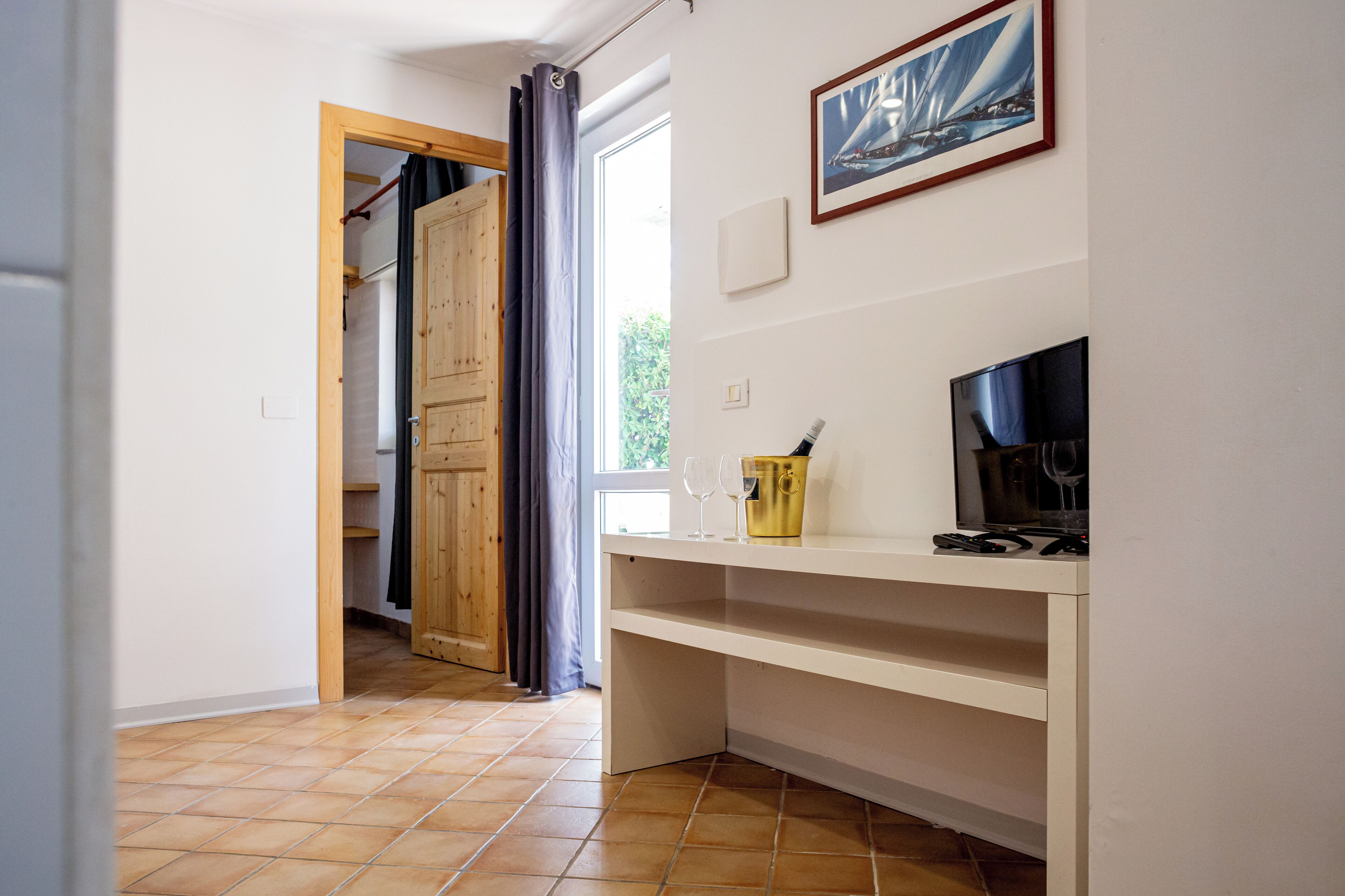 Una habitación con puerta de madera, ventana con cortinas, televisor y una pequeña mesa con una copa de vino y una botella.