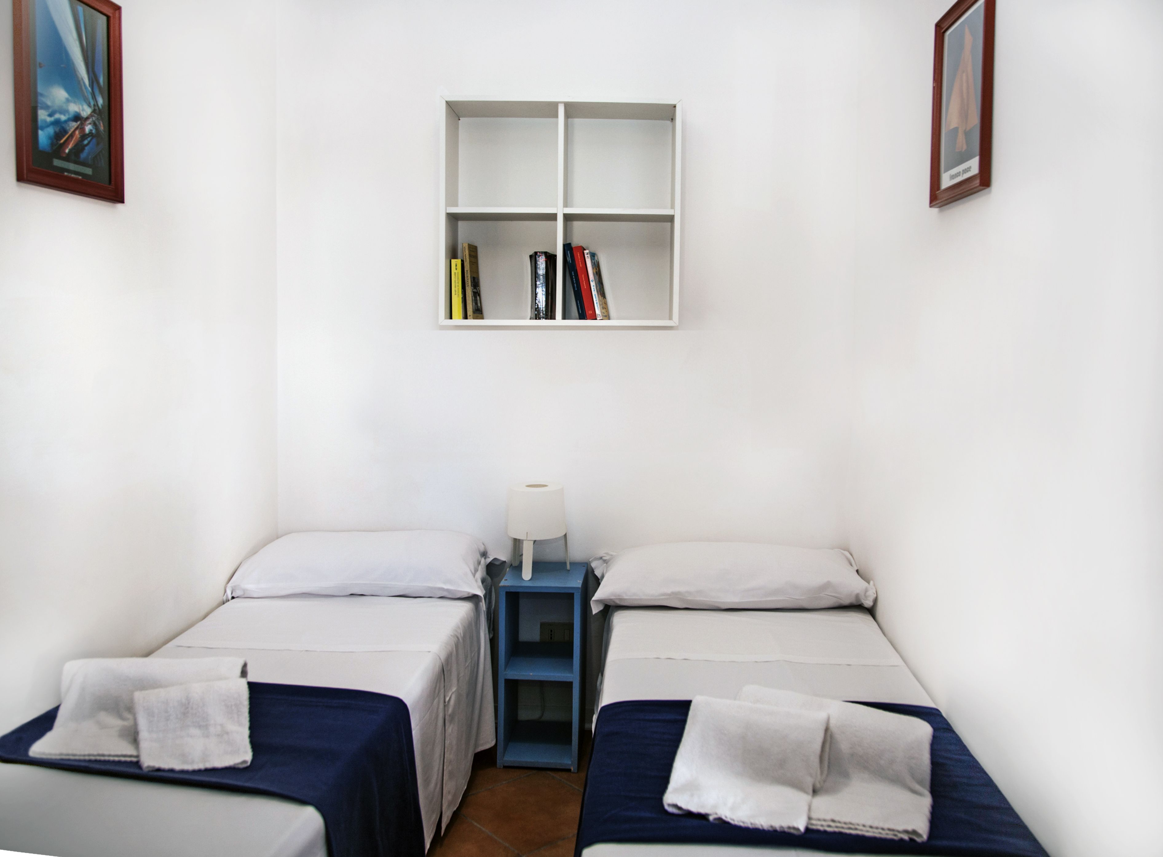 Ein Zimmer mit zwei Betten, einem kleinen Tisch mit Lampe und einem Bücherregal mit Büchern.