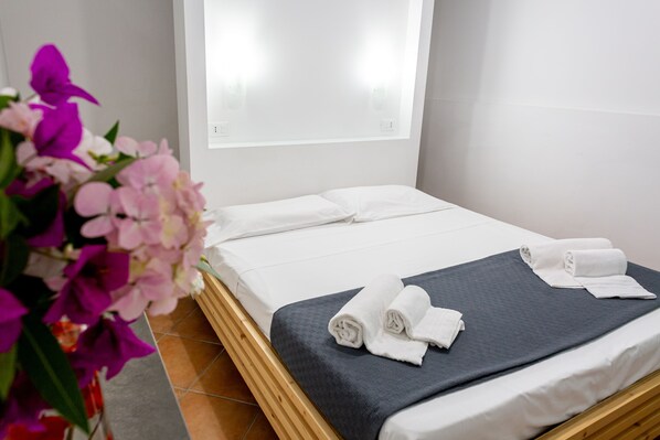 A neatly made bed with white linens and a grey blanket, a wooden headboard, and a vase of pink flowers on the floor. - Residence "Il Cicero" (Gioiosa Marea)