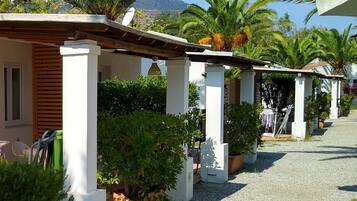 A row of white-pillared buildings with a gravel pathway, surrounded by lush greenery and palm trees.