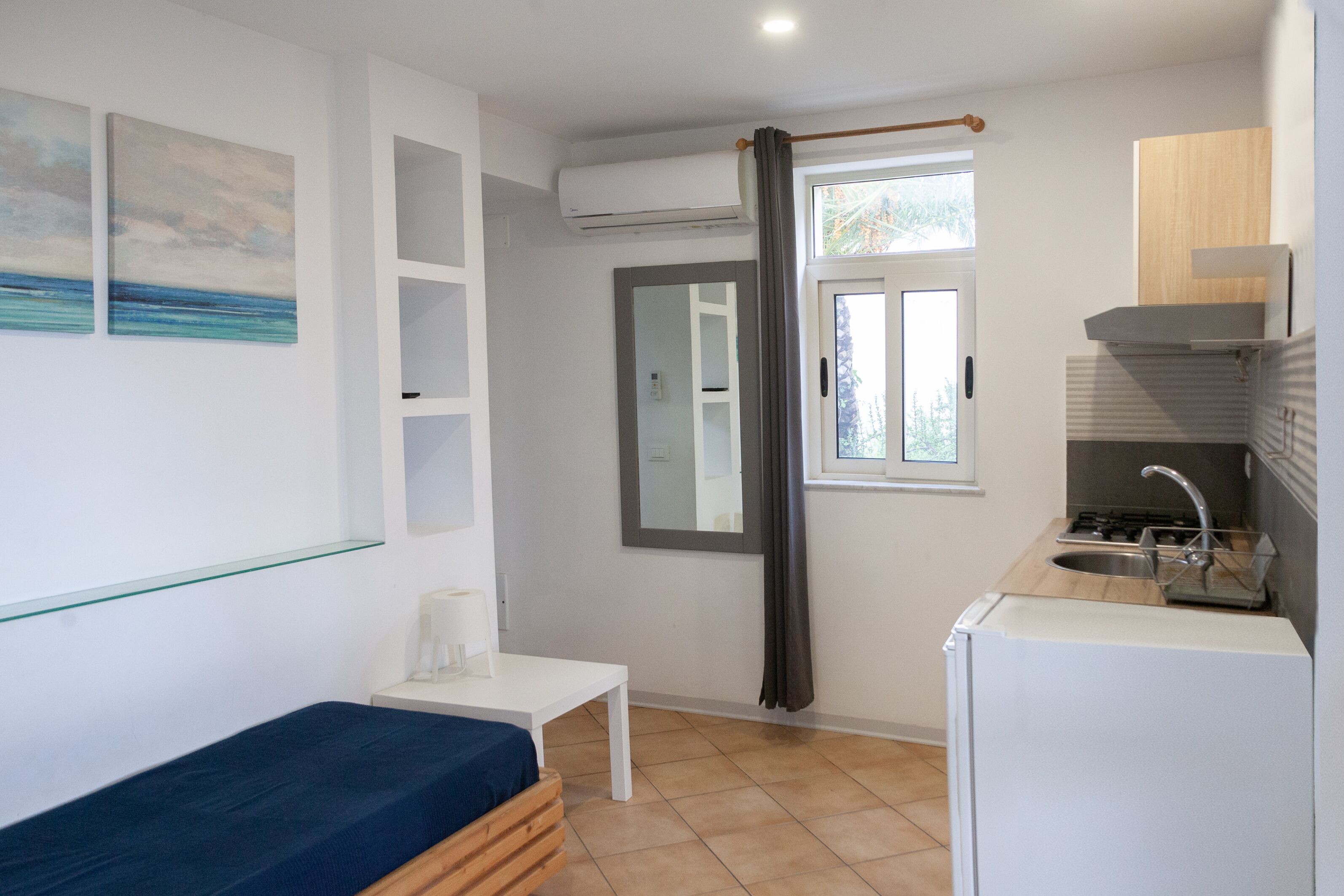 A compact room with a bed, a small table, a window with a view, a kitchenette, and a mirror.