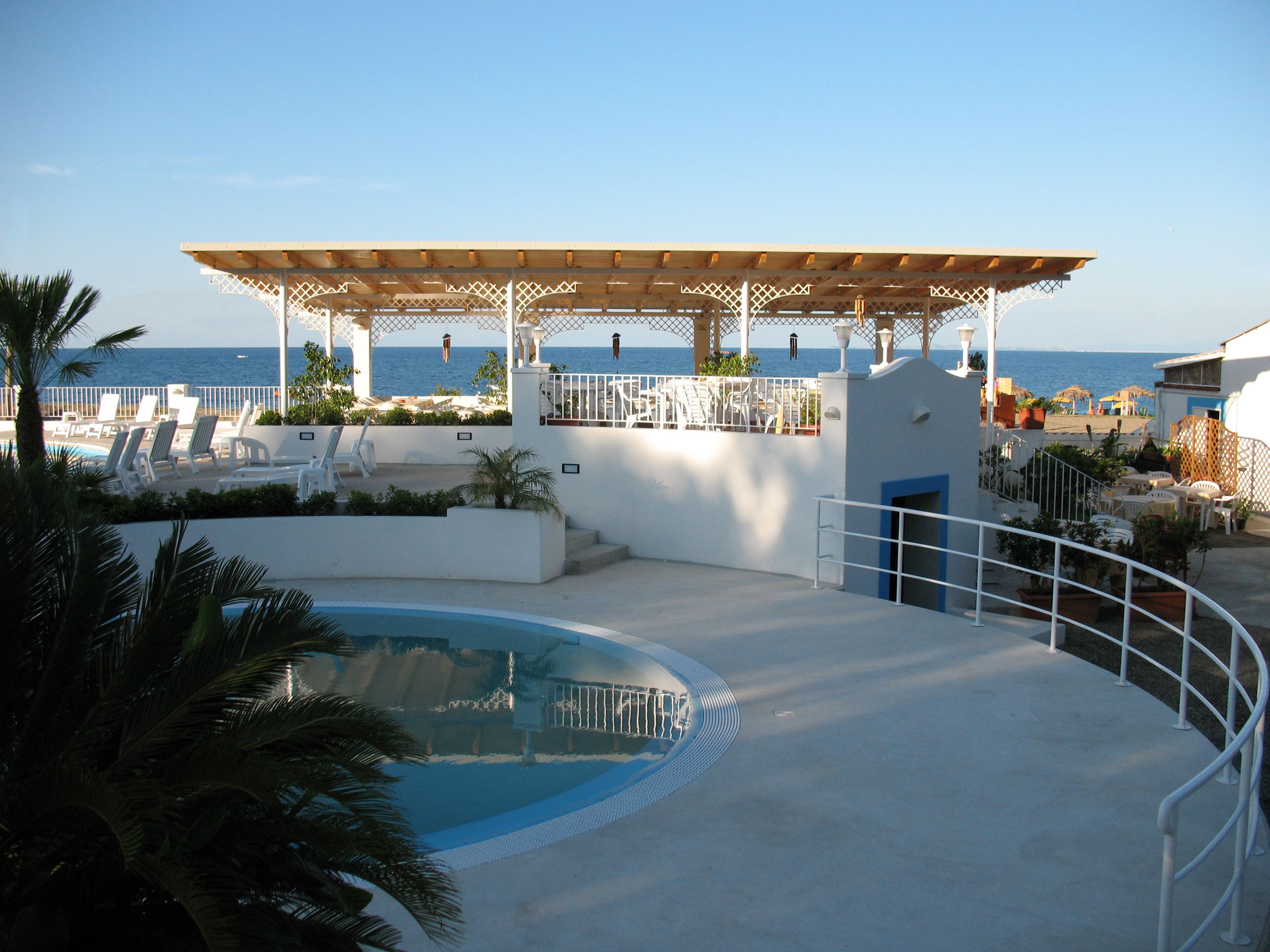 A pool area with lounge chairs, a pergola, and a view of the sea.