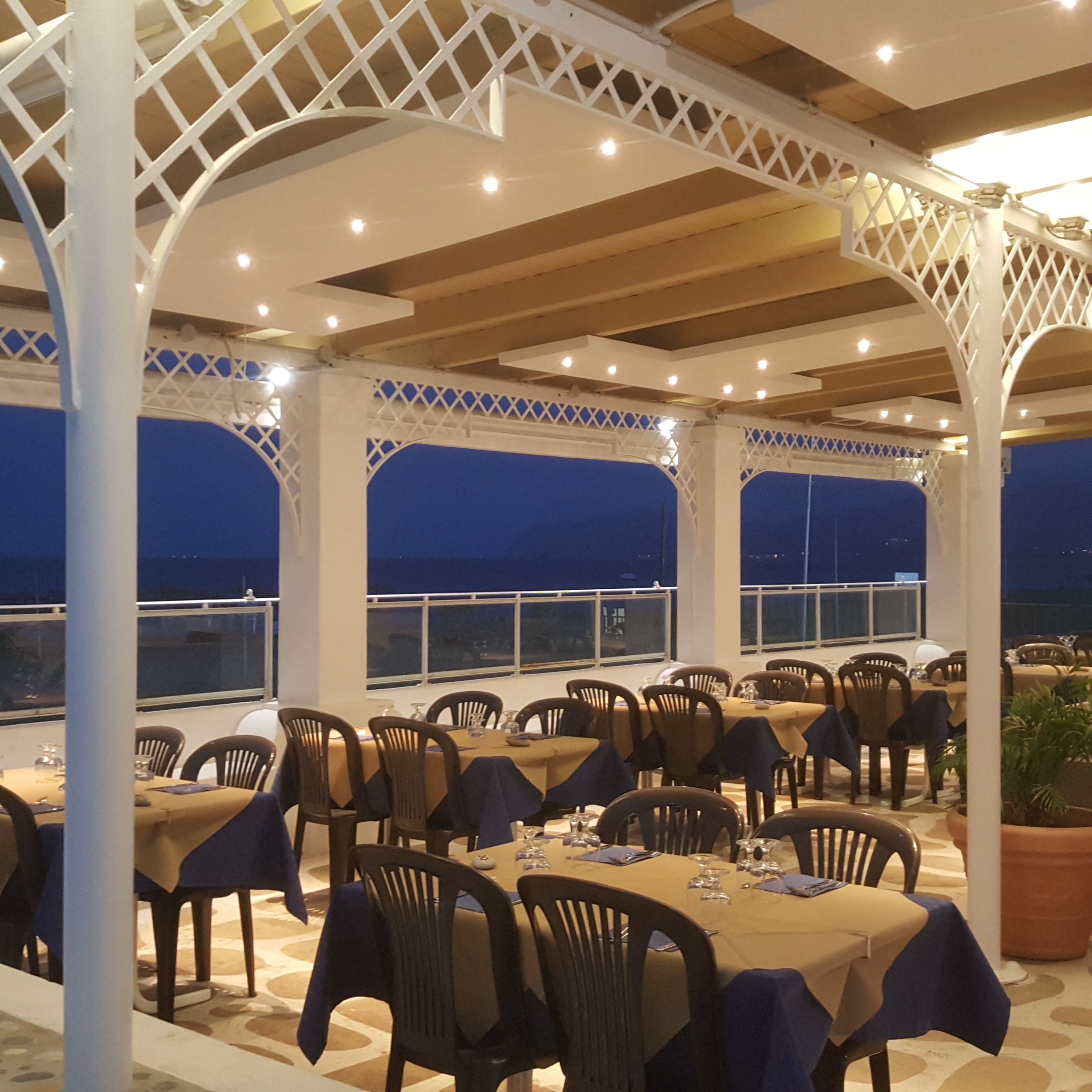 Un área de comedor al aire libre con mesas y sillas, cubierta por una estructura de celosía blanca, y ventanas amplias con vistas al mar.