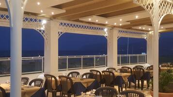 Un área de comedor al aire libre con mesas y sillas, cubierta por una estructura de celosía blanca, y ventanas amplias con vistas al mar.