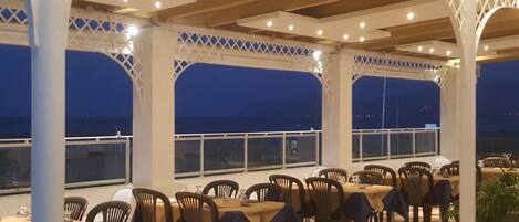 An outdoor dining area with tables and chairs, covered by a white lattice structure, and large windows offering a view of the sea.