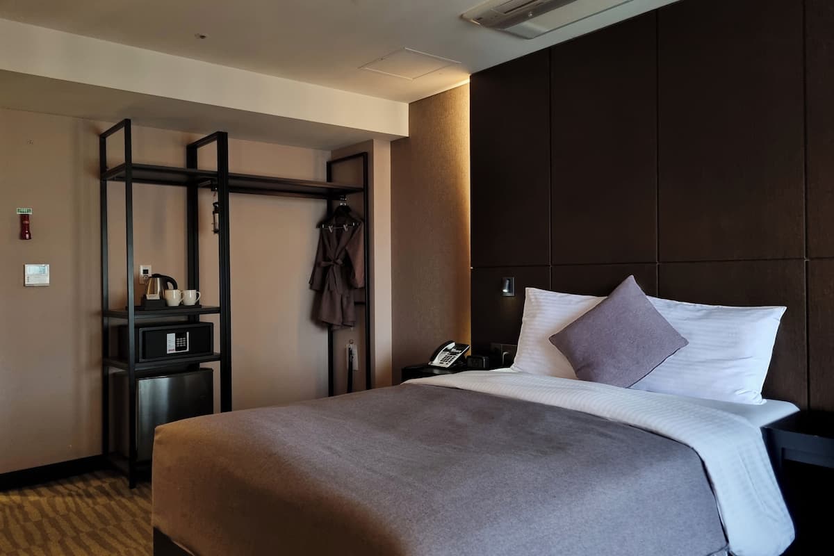 Une chambre d’hôtel avec un grand lit, une table de chevet avec un téléphone, un peignoir suspendu à un porte-manteau et un panneau de commande au mur.