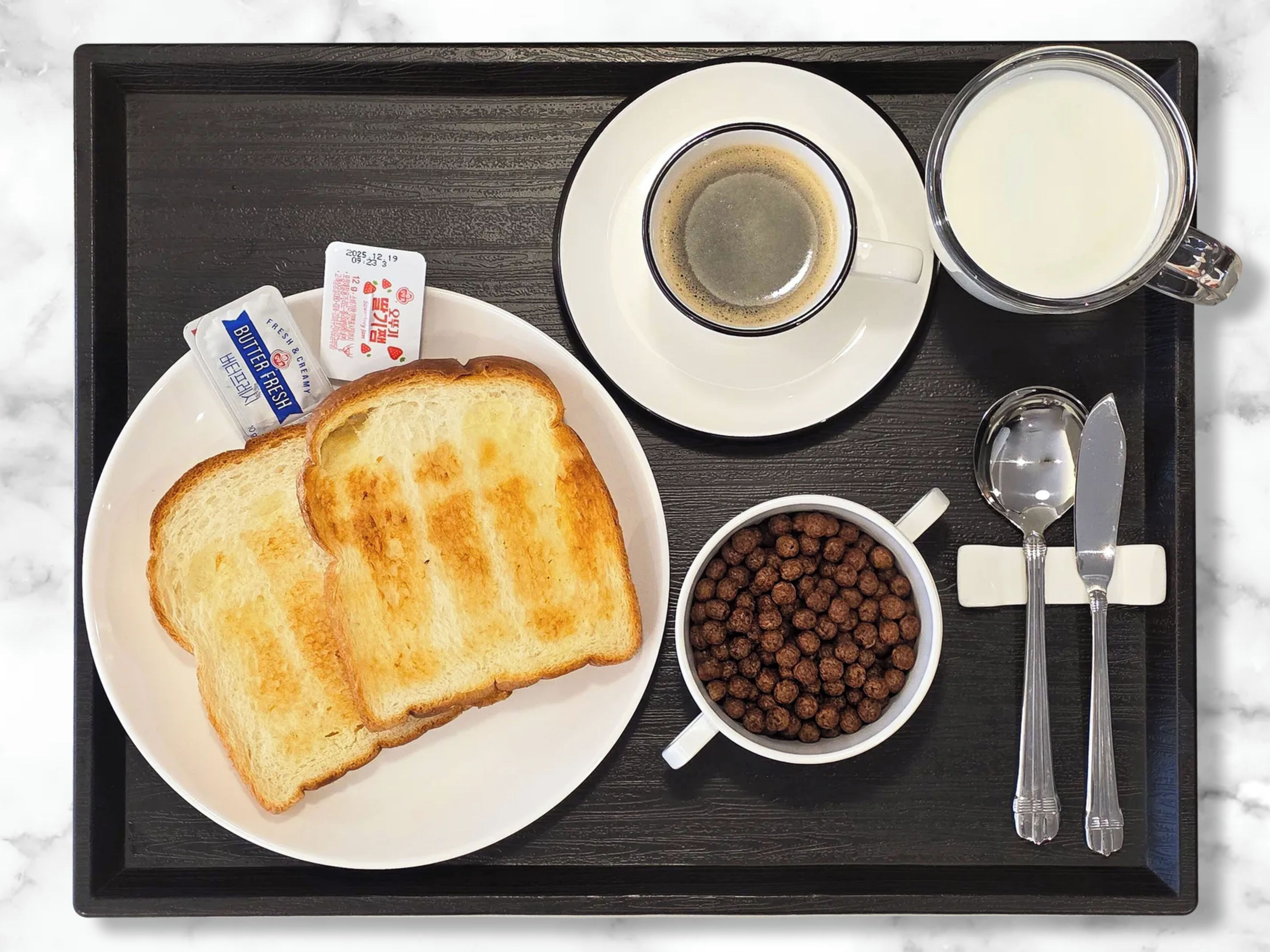 A tray with toast, a cup of coffee, a mug of milk, a bowl of chocolate cereal, and a packet of butter & cream.