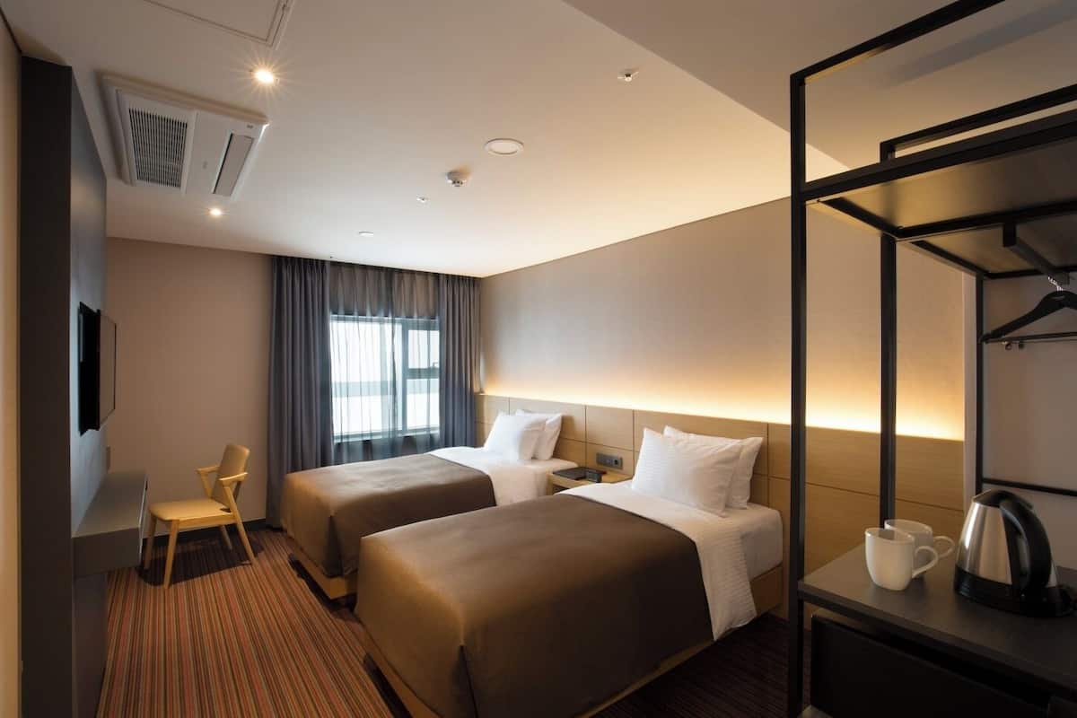 Une chambre d’hôtel avec deux lits, un bureau avec une bouilloire, une chaise et un téléviseur fixé au mur.