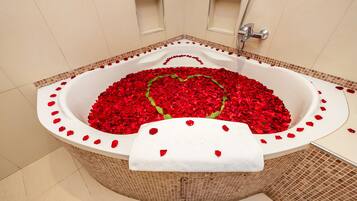 Una bañera llena de pétalos de rosa roja formando un corazón, con una toalla blanca colgada al borde.