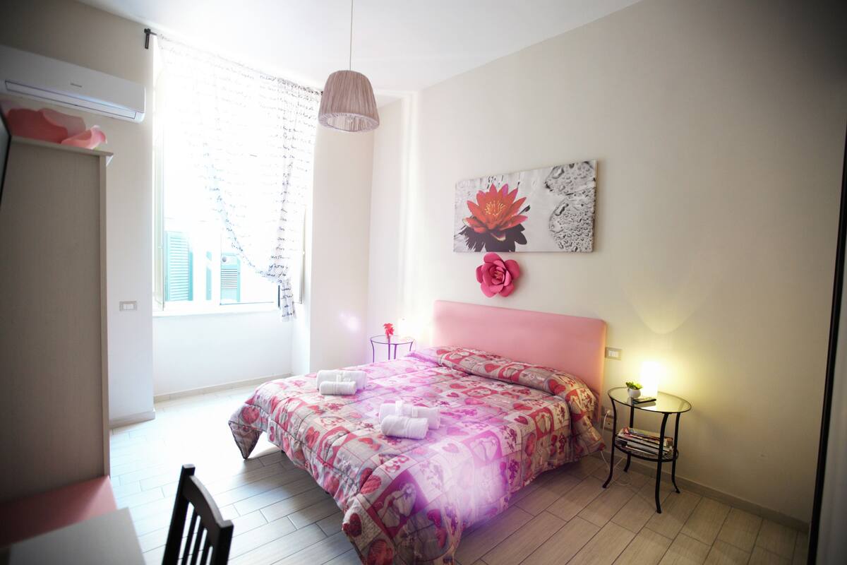 A bedroom with a bed, a bedside table with a lamp, a window with blinds, and a wall with a floral painting.