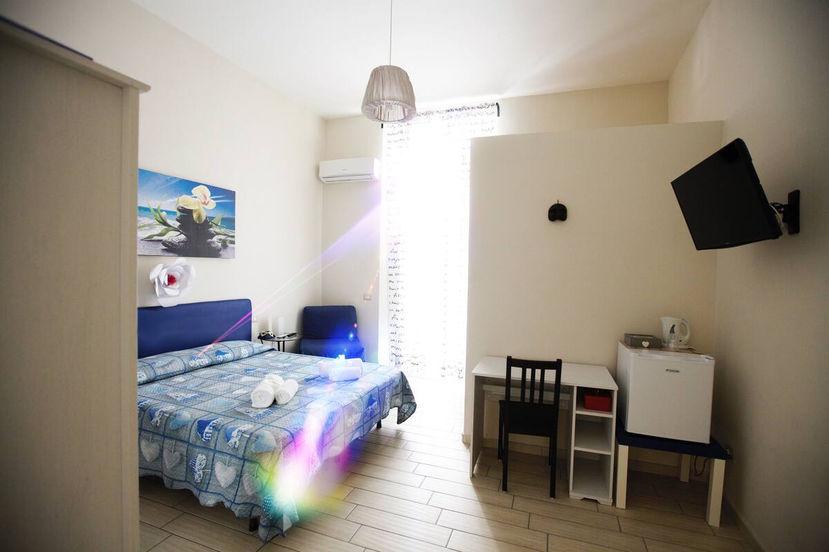 A bedroom with a bed, a desk, a chair, a TV, and a window with curtains.