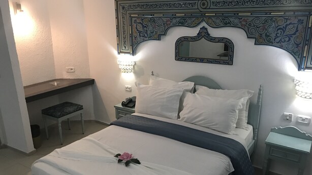 Una cama bien hecha con sábanas blancas, un espejo decorativo encima y una mesita de noche con una lámpara.