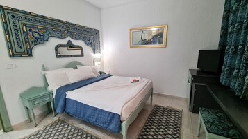 Un dormitorio con cama, mesita de noche, cómoda, televisor, alfombras estampadas y decoración en la pared.