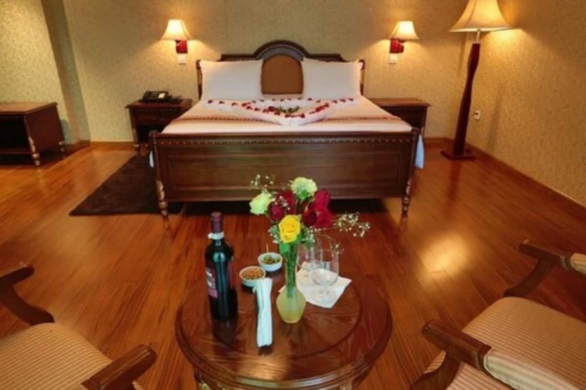 A hotel room with a wooden floor, a bed with a white headboard, two bedside tables, a round glass table with a bottle, a vase with flowers, and two chairs.