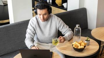 A person using a laptop on a grey couch with a small table beside them containing a bowl of croissants, a glass bottle, and a potted plant.