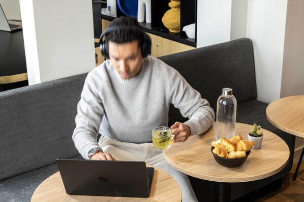 Una persona usando una laptop en un sofá gris con una mesita auxiliar cercana que tiene un bol de croissants, una botella de vidrio y una planta en maceta.
