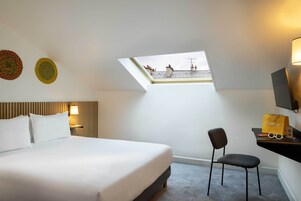 Une chambre d’hôtel avec un grand lit, une lucarne, une chaise, un bureau avec une lampe et un téléviseur.