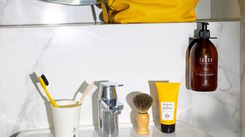 Um banheiro com parede de mármore, uma bolsa amarela, um frasco de gel Terre Mars, uma escova de barbear e uma escova de dentes em um copo.