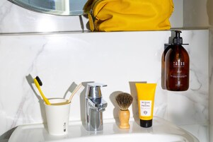 A bathroom with a marble wall, a yellow bag, a bottle of Terre Mars gel, a shaving brush, and a toothbrush in a cup.