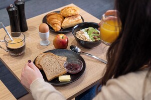 A breakfast table with croissants, toast, jam, butter, a glass of orange juice, a bowl of cereal, a glass of coffee, and a plate with an apple.
