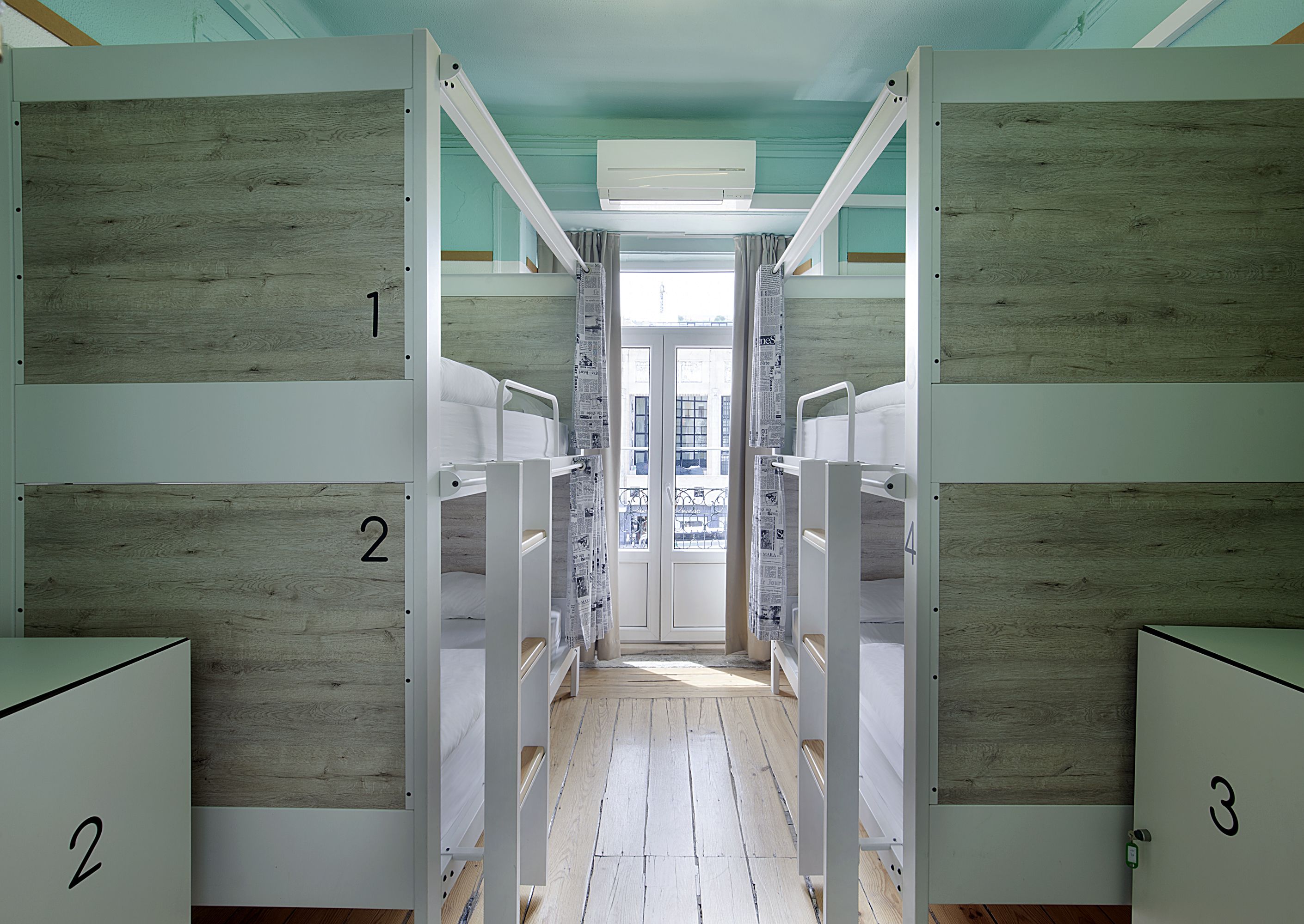 A corridor with bunk beds numbered 1 to 4, wooden walls, and a window with a view of buildings.