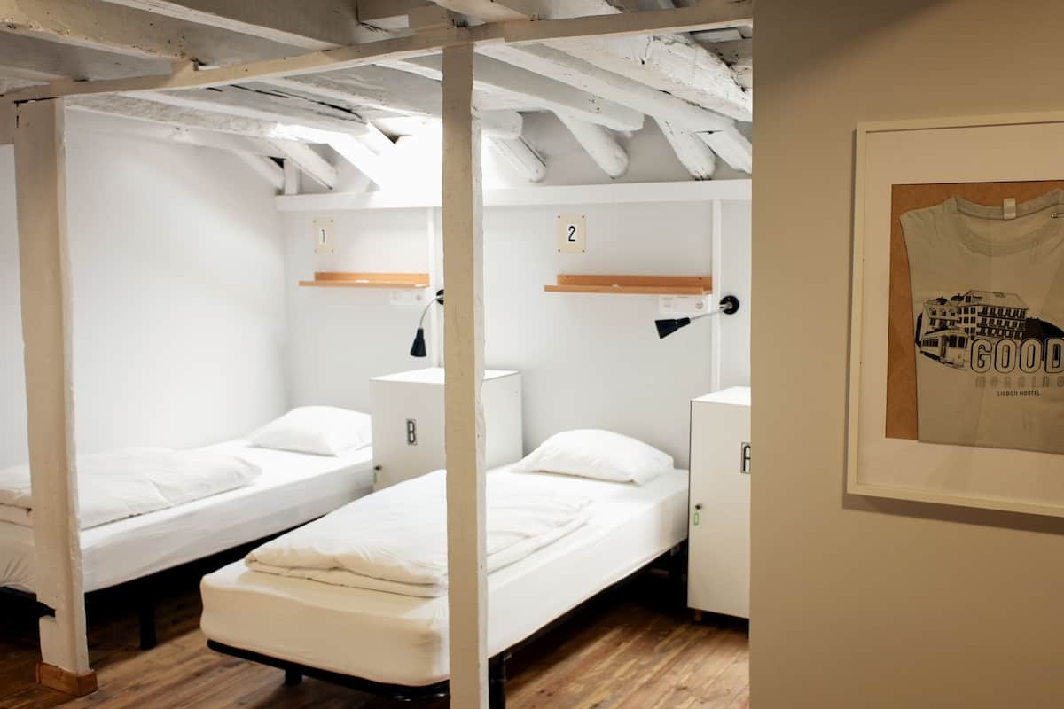A dormitory room with two single beds, wooden beams on the ceiling, a small desk, and a framed poster on the wall.