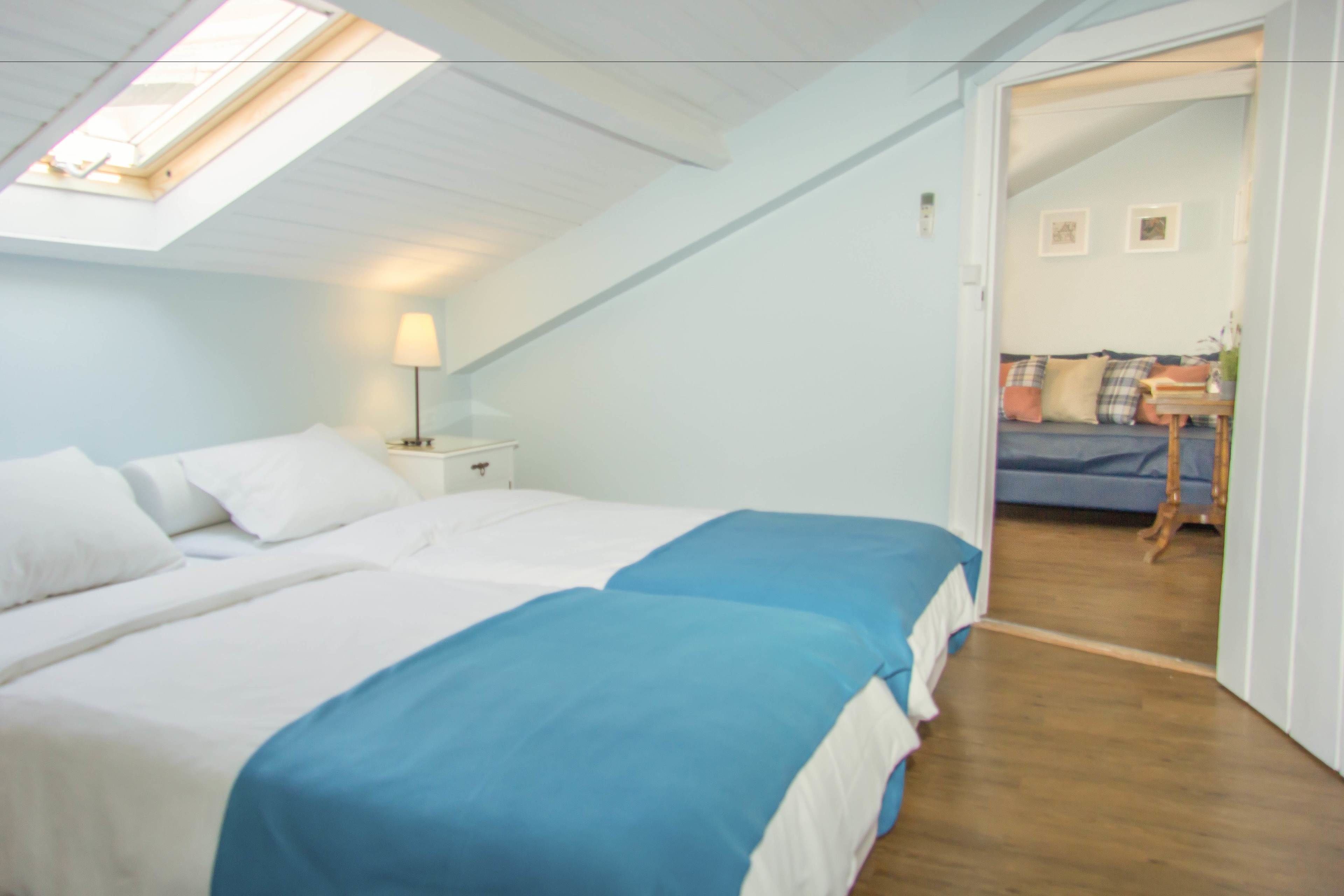 A bedroom with two beds, a skylight, a bedside table with a lamp, and a view into another room with a sofa and a table.