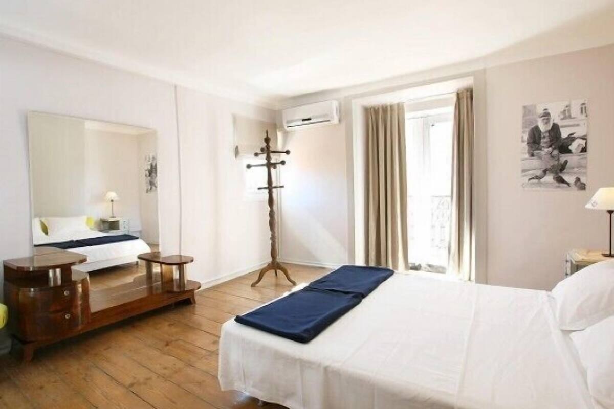 A hotel room with a bed, a wooden floor, a mirror, a coat stand, a window with curtains, and a wall-mounted air conditioner.