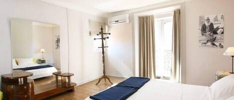A hotel room with a bed, a wooden floor, a mirror, a coat stand, a window with curtains, and a wall-mounted air conditioner.
