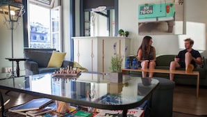 A modern living room with a glass coffee table, books, and magazines. There is a chessboard on the table, and a green sofa with cushions. A potted plant is on the table, and a car is parked outside.