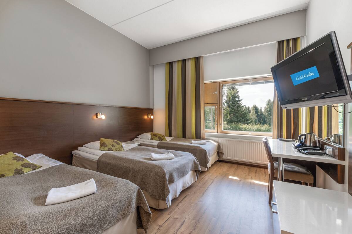 Habitación de hotel con dos camas, un escritorio con televisor y ventana con vista a árboles.
